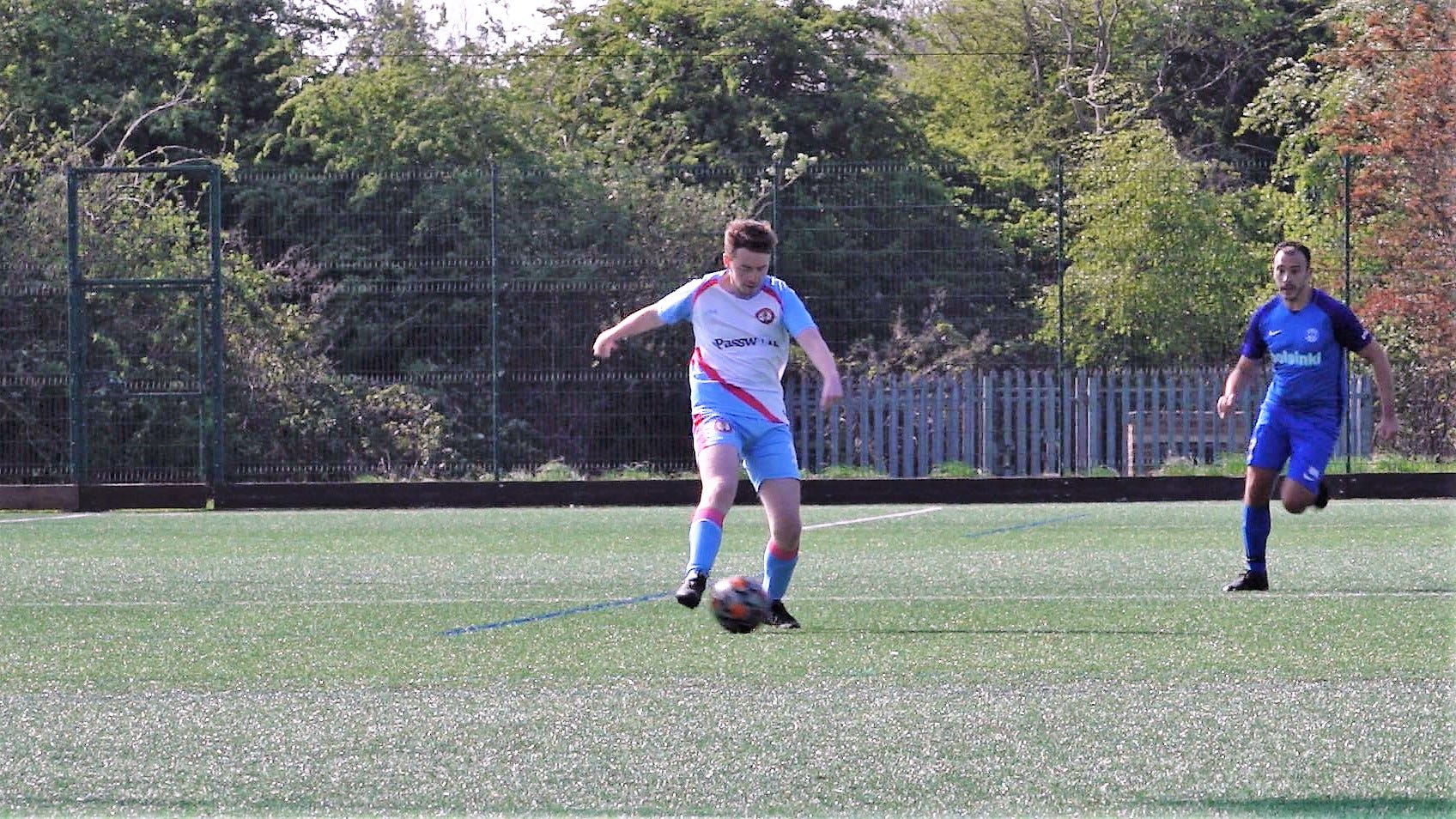 A man in a white, pink, and blue kit runs with the ball. Behind him, a man in a darker blue kit chases.