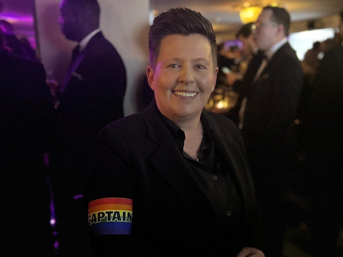 Woman with short dark brown hair smiles and wears a black suit. On her arm, there's a rainbow captain's armband.