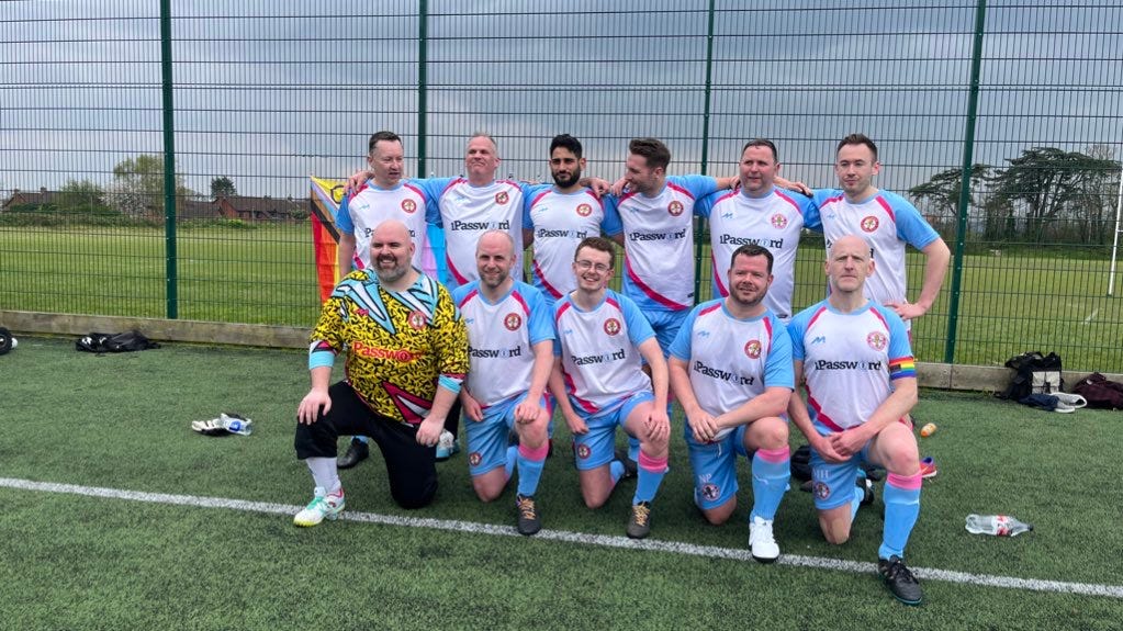 A team photo of 11 masculine-presenting people in two rows on an astroturf pitch. Behind them, there's a small pride progress flag.