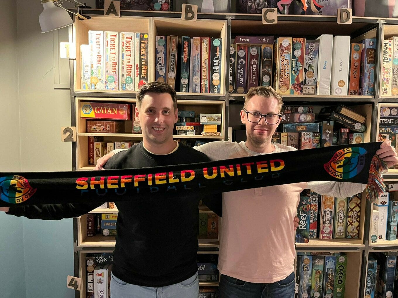 In front of shelves full of board games, two men hold up a scarf showing the words “Sheffield United” in rainbow colours