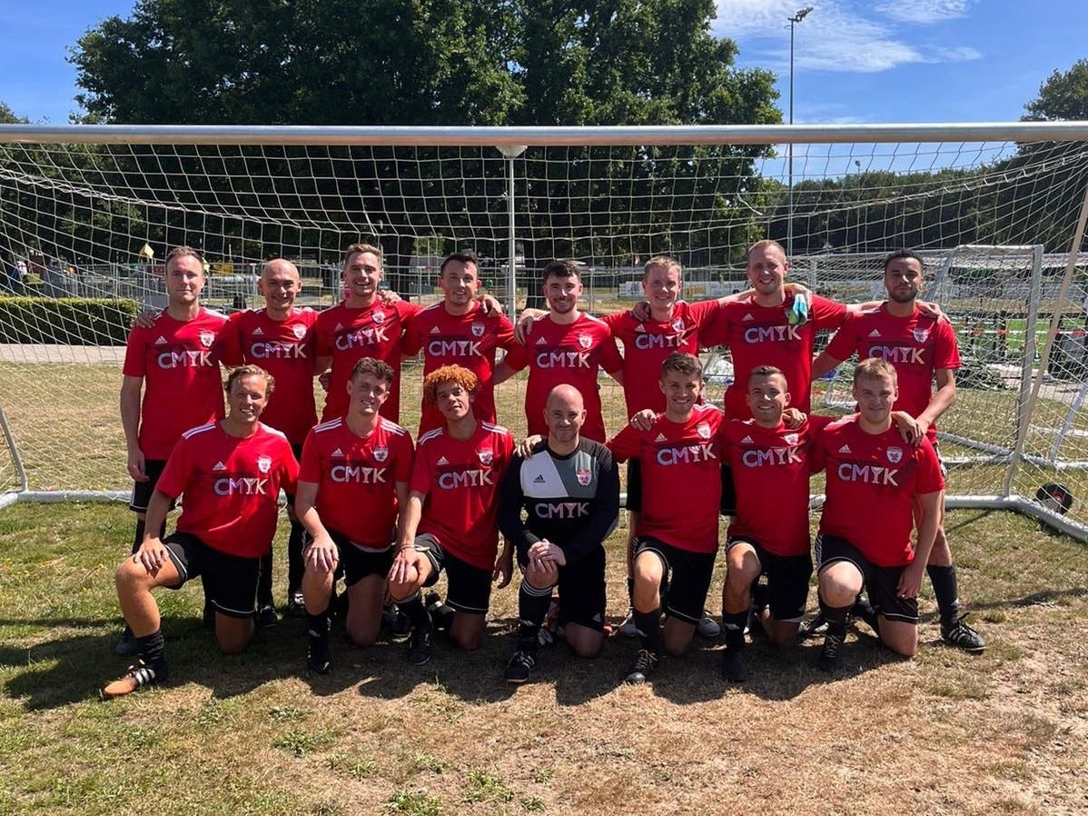 A football squad photo. A group of 15 men in red football kits are in two rows, one row standing and the other kneeling in front. They're in front of a football goal and smiling.