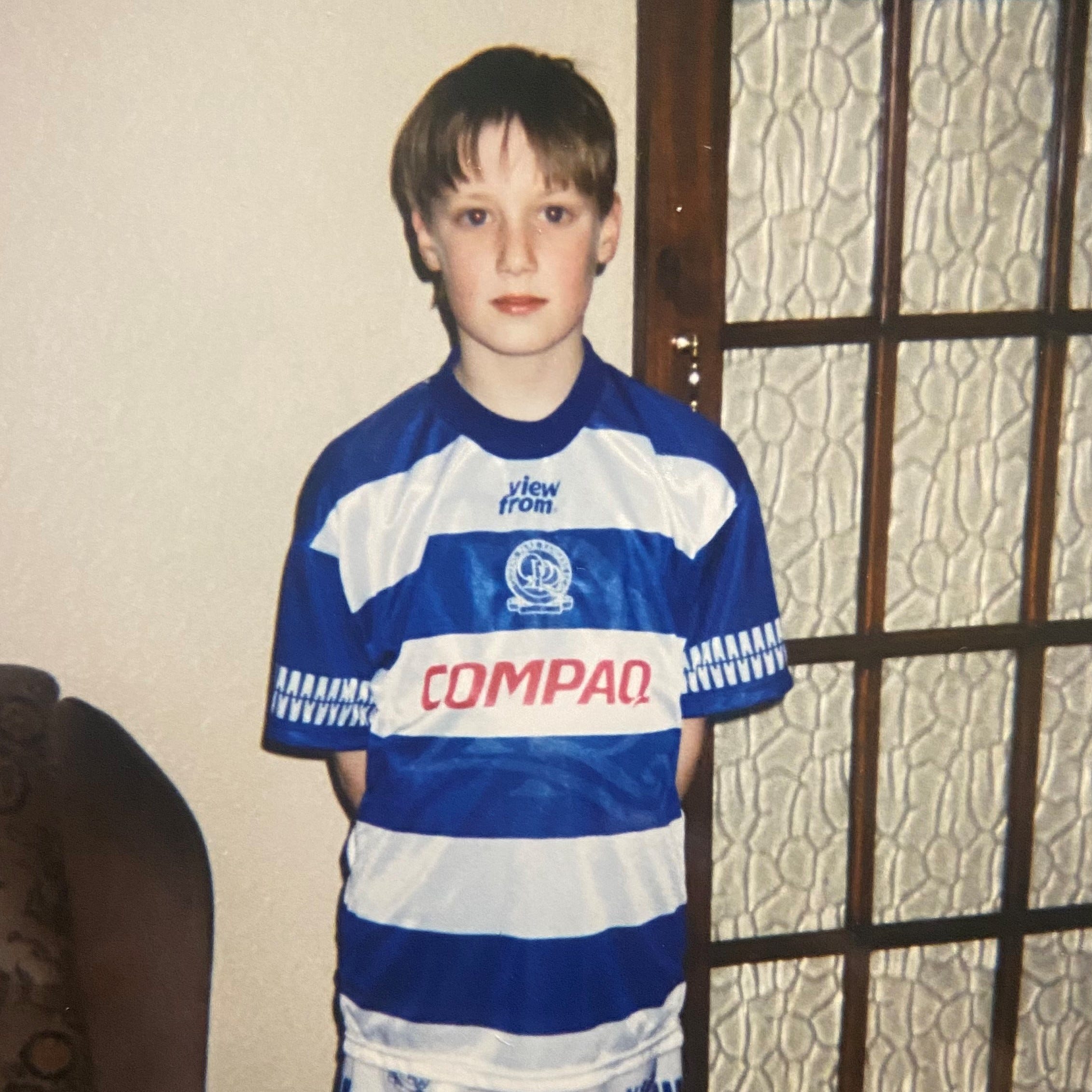 A young man with 90s-style curtains is standing against a wall and looking at the camera, wearing a QPR shirt with large horizontal blue and white stripes