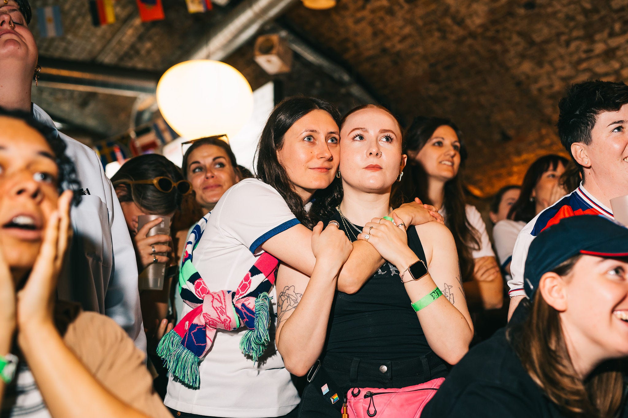 Among a crowd of people, two female-presenting people look on while embracing