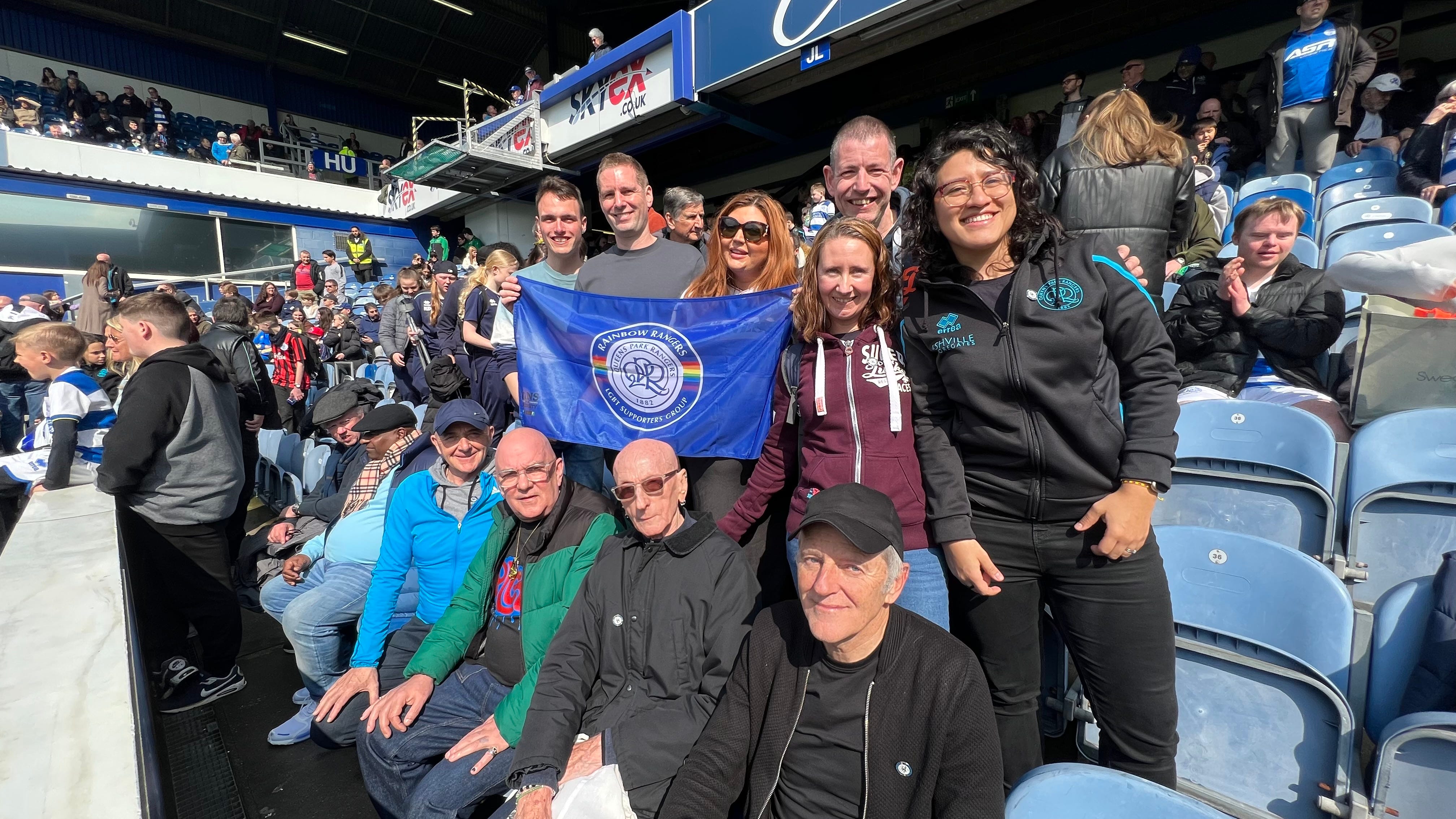 Two rows of people at their seats at Loftus Road football stadium. The front row is sitting, and the back one is standing. They're all looking at the camera and smiling. On the back row, a flag that reads "Rainbow Rangers LGBT supporters group" is being held up.