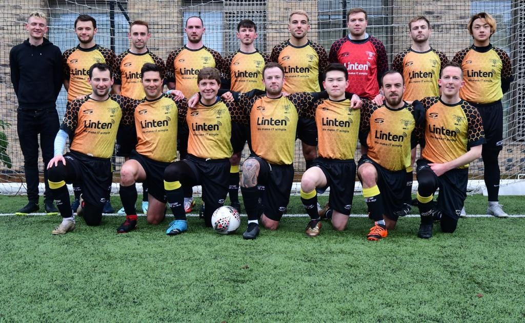 A football squad pose on a football pitch in two lines, wearing yellow football shirts and black shorts. A man with blonde hair is standing on the second row to the left.