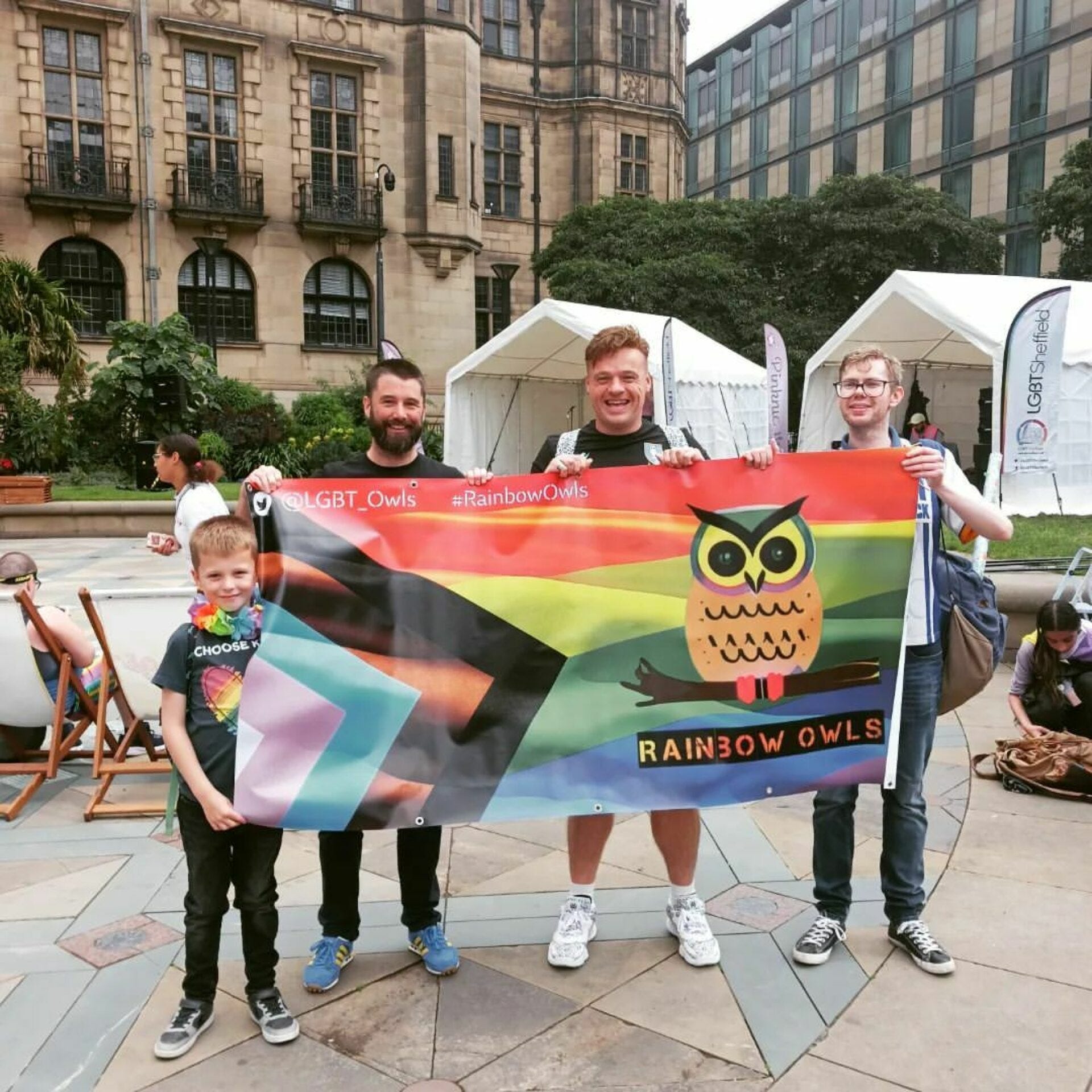 Three men and a boy hold up a Pride Progress flag with the rainbow owls logo printed on it.