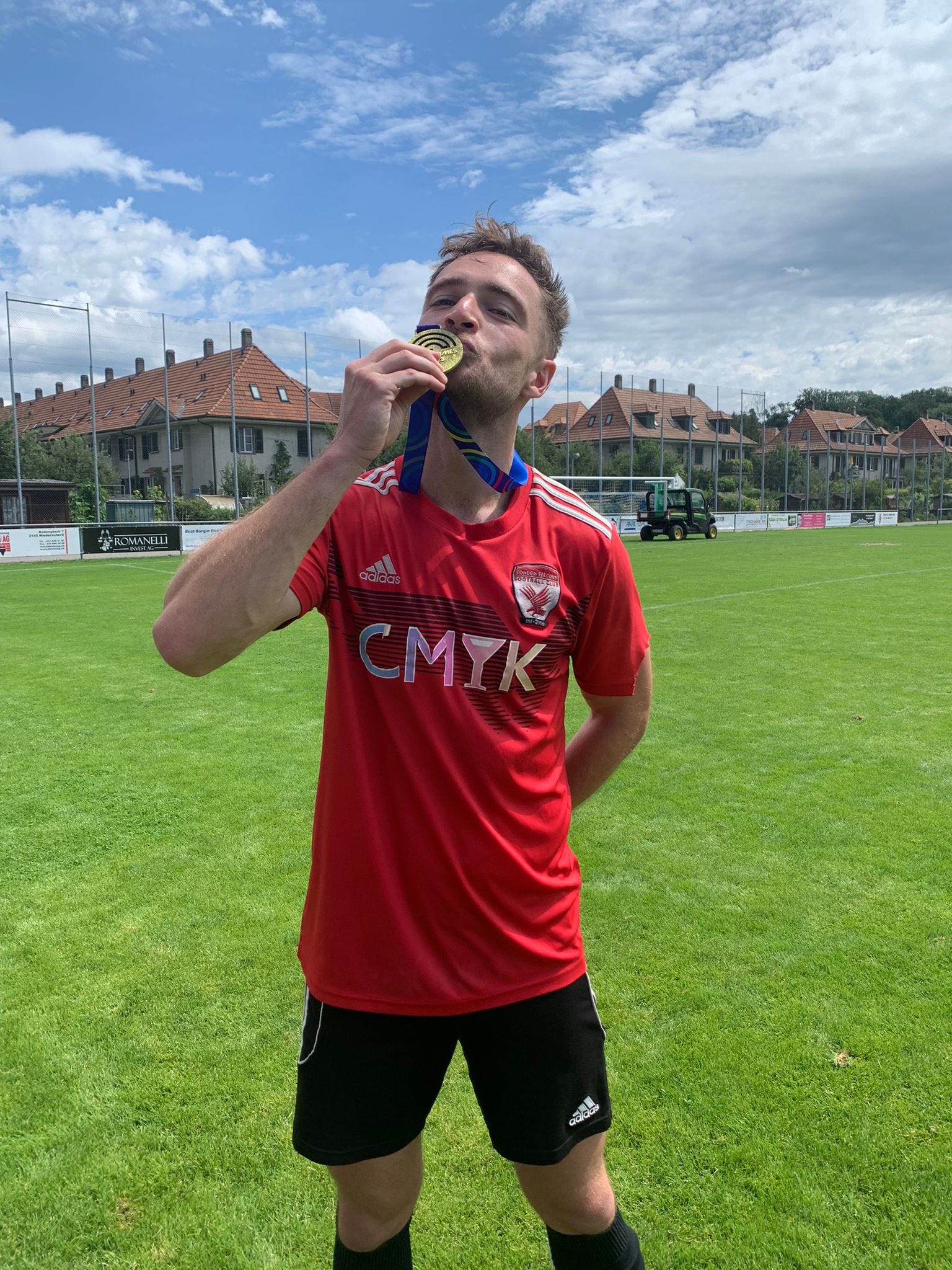 On a football pitch, Will - a white man with short brown hair - kisses his medal. He's wearing a red adidas football shirt with a falcon on the crest and black shorts.