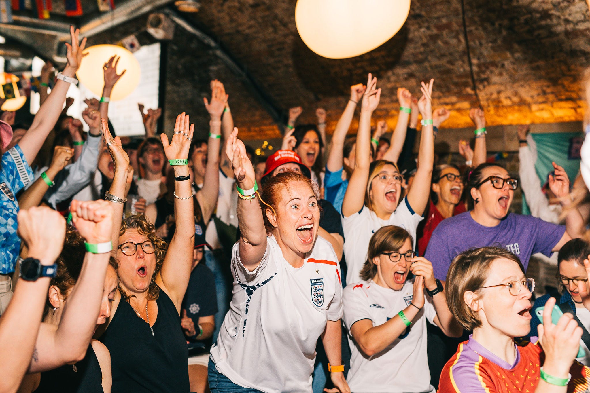 In a railway arches bar, a crowd of mostly female-presenting people celebrate, wearing a variety of football shirts