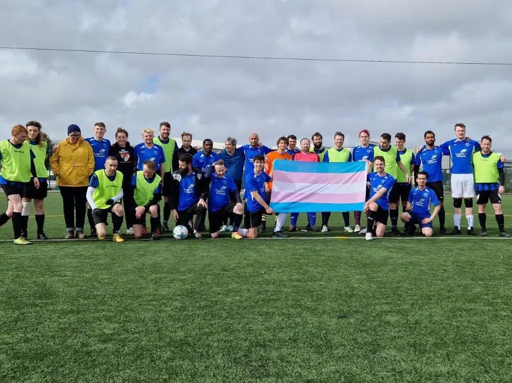 A large group of people on a football pitch hold up a trans flag