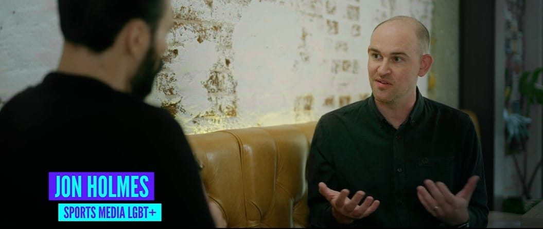 Screenshot of a documentary interview. Jon Holmes, a man with a dark green button-up shirt holds his hands out as he speaks to Rylan. The lower third caption reads "Jon Holmes, Sports Media LGBT+"