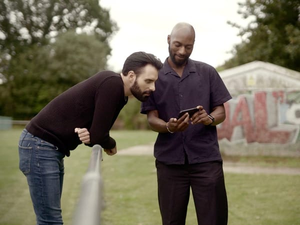 Two men stand on a recreational ground, one leaning over to watch something on a phone in the other's hands.