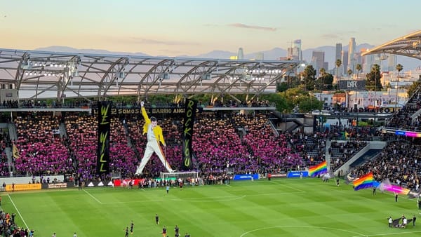 LA soccer stadium at sunset, a huge banner of Freddie Mercury with his fist clenched drapes down from the roof of a stand